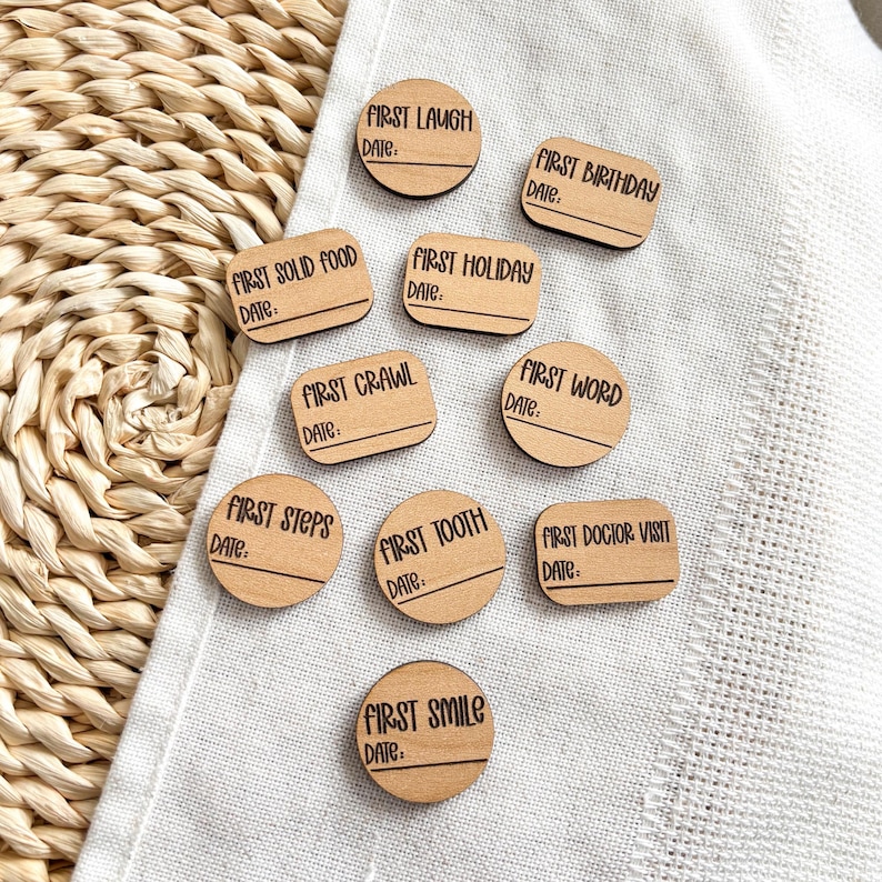 Puede incluir: Un conjunto de marcadores de hitos de madera con texto negro sobre tela blanca. Los marcadores tienen varias formas, incluyendo c&iacute;rculos y rect&aacute;ngulos. Cada marcador tiene una frase como "Primera risa" o "Primer cumplea&ntilde;os" y una l&iacute;nea para la fecha.