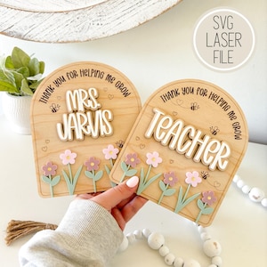 Peut inclure: Deux plaques en bois pour remercier les enseignants. L'une dit "Mrs. Jarvis" et l'autre "Teacher" en lettres blanches superposées. Les deux ont un motif floral avec des fleurs roses et violettes, des tiges vertes et de petites abeilles. Le texte "Thank you for helping me grow" est arqué au-dessus.