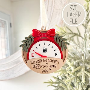 May include: A round wooden Christmas ornament featuring a fuel gauge design with a red needle pointing to E. It includes a red bow, green leaf accents, and the text "THE YEAR WE COULDN'T afford gas 2026" engraved on the front, suspended by a white ribbon.