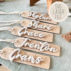 Op de afbeelding: Houten naamlabels met witte, verhoogde letters die namen spellen als Skylar, Shari, Grace, Trenton en Grams. Elk label heeft een feestelijk patroon van kerstbomen en cadeaus. De labels zijn bevestigd aan een natuurlijk touw.