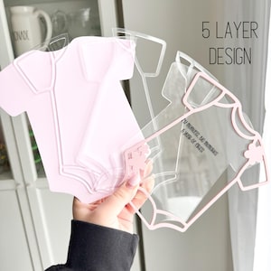 Puede incluir: Un dise&ntilde;o de body de beb&eacute; multicapa en acr&iacute;lico rosa y transparente. El texto "5 LAYER DESIGN" es visible. El dise&ntilde;o incluye el texto "My moments, big memories, A year of firsts."