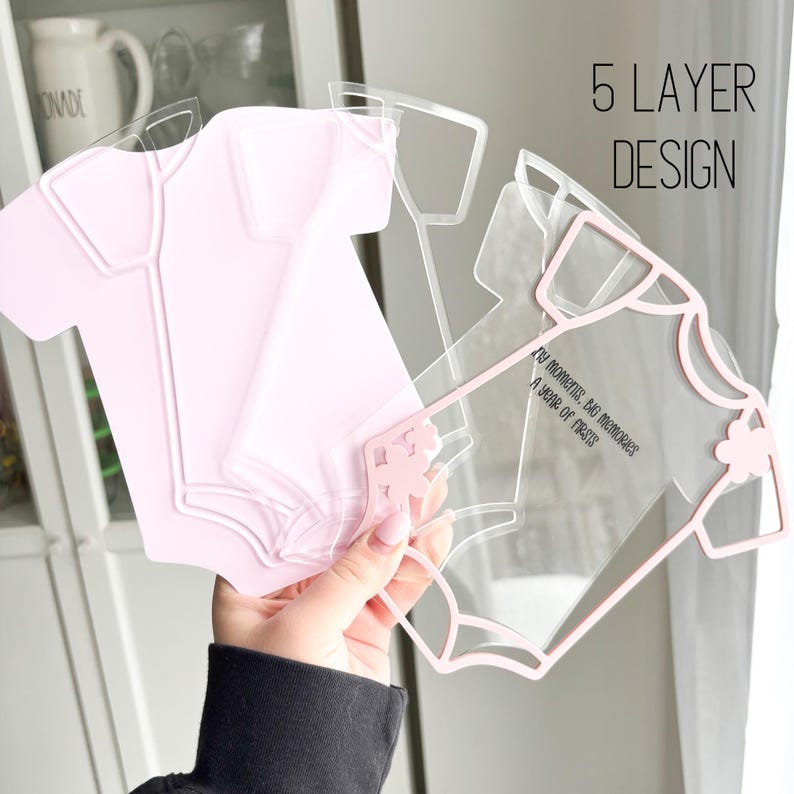 Puede incluir: Un dise&ntilde;o de body de beb&eacute; multicapa en acr&iacute;lico rosa y transparente. El texto "5 LAYER DESIGN" es visible. El dise&ntilde;o incluye el texto "My moments, big memories, A year of firsts."