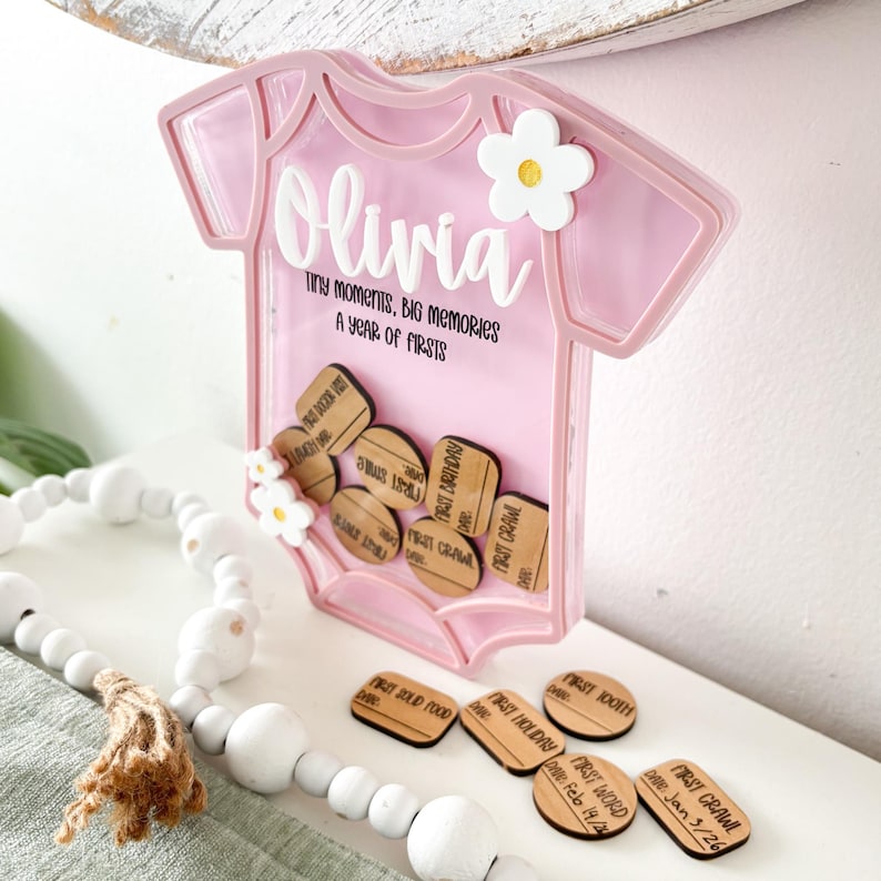 Puede incluir: Una caja de recuerdos rosa con forma de body de beb&eacute; con el nombre "Olivia" en escritura blanca. Incluye fichas de madera para registrar los primeros hitos del beb&eacute;. Tambi&eacute;n se ve el texto "Tiny moments, big memories, a year of firsts".