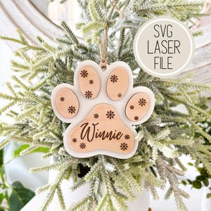 May include: White wooden paw print ornament with the name "Winnie" engraved on it. The ornament is decorated with snowflakes and is suitable for a Christmas tree.