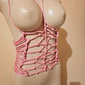 Pode incluir: Top de macramé rosa feito à mão. O top apresenta um decote halter e um design tecido no peito e no torso. O macramé é amarrado para criar um padrão semelhante a uma treliça. A peça de roupa é exibida em um manequim.
