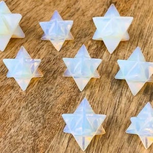 May include: Eight opalescent star-shaped crystals are arranged on a wooden surface. The crystals have a six-pointed star design with a translucent, iridescent quality, reflecting light with a soft glow. The crystals vary slightly in size and are evenly spaced.