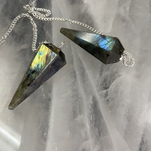 Op de afbeelding: Twee labradoriet kristallen pendels met zilveren kettingen. De pendels zijn facetgeslepen, met een puntig uiteinde en een zilveren lus aan de bovenkant. De kristallen vertonen iriserende flitsen van blauw, groen en geel. De achtergrond is een wit, doorschijnend oppervlak.