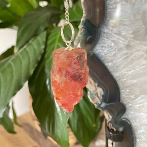 May include: A red carnelian crystal pendulum on a silver chain. The crystal is a raw, natural shape with a pointed tip. The chain has a silver ring at the top.