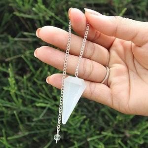 Könnte beinhalten: Ein weißer Kristallpendel mit einer silbernen Kette. Der Pendel hängt an der Hand einer Person.