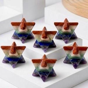May include: Six star-shaped decorative objects. Each star features a layered design with colours of the rainbow, including red, orange, yellow, green, blue, and purple. The top portion is a reddish-brown colour. The objects are arranged on a white surface.