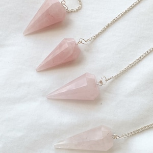 May include: Four pink rose quartz crystal pendulums with silver chains. The pendulums are all the same shape and size, and they are hanging from the chains.