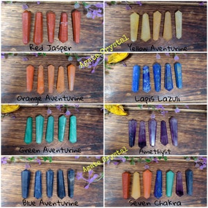 May include: Seven chakra crystal wands arranged in a grid. The wands are red, orange, yellow, green, blue, indigo, and violet. The wands are displayed on a wooden surface. The text "Agate Crystal" is written on the image.