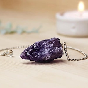 Op de afbeelding: Een paarse amethist kristallen slinger aan een zilveren ketting. De kristal is ruw en heeft een natuurlijke vorm.