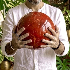 Puede incluir: Una gran piedra de jaspe rojo esférica y pulida con vetas naturales visibles en tonos naranja y crema. La superficie es lisa y reflectante, mostrando una paleta de colores carmesí profundo con patrones orgánicos intrincados en la esfera.