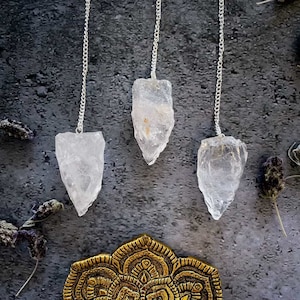 May include: Three clear quartz crystal pendulums hanging from silver chains against a grey textured background. The pendulums are all different shapes and sizes.