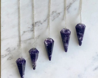 Pode incluir: Cinco pêndulos de cristal de ametista com correntes prateadas penduradas em uma superfície branca. Os pêndulos são todos pontiagudos na parte inferior e têm um padrão de mármore roxo e branco.