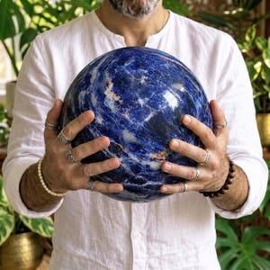 Puede incluir: Una gran esfera de sodalita pulida que presenta una paleta de colores azul profundo con vetas blancas y grises prominentes en toda su superficie. La piedra es lisa, redondeada y muestra patrones minerales naturales en todo su exterior.
