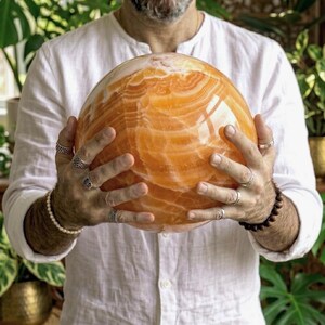 以下が含まれることがあります： 琥珀色、金色、クリーム色の自然な縞模様と渦巻き模様が見える、大きく磨かれたオレンジ色のカルサイトの球体。表面は滑らかで光沢があり、球体全体に半透明の鉱物層が広がっている様子が確認できます。