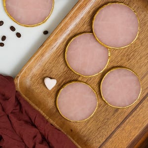 May include: Five pink, circular coasters with gold edging, presented on a wooden tray. A small white heart-shaped item and coffee beans are also visible. A maroon cloth is draped in the corner of the image.
