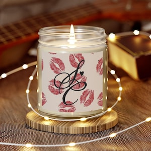 May include: A lit candle in a clear glass jar with a wooden coaster. The jar has a label with red lipstick kiss marks and a black cursive "J" with a heart. The candle is surrounded by a string of small lights.