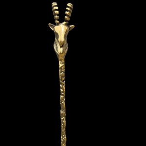 May include: A gold-toned metal hair stick featuring a sculpted antelope head at the top with two ridged, curved horns. The long, slender shaft of the hair accessory displays a textured, organic pattern along its entire length, finished in a polished metallic sheen.