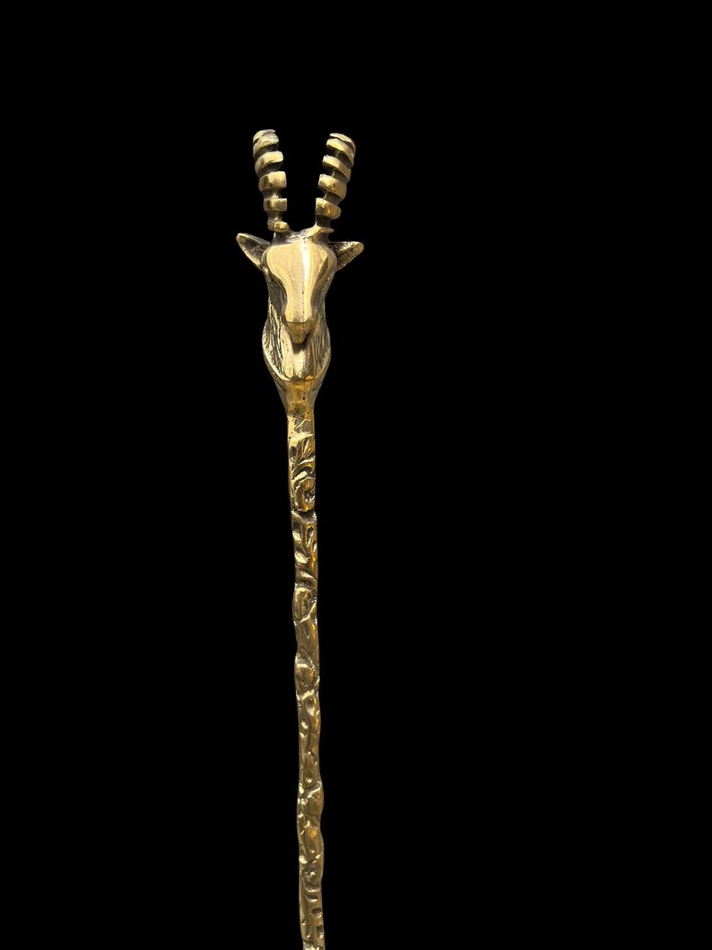 May include: A gold-toned metal hair stick featuring a sculpted antelope head at the top with two ridged, curved horns. The long, slender shaft of the hair accessory displays a textured, organic pattern along its entire length, finished in a polished metallic sheen.
