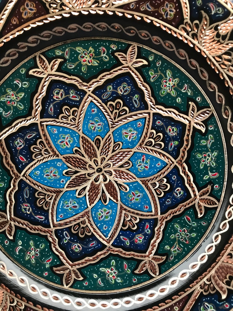 Puede incluir: Un plato decorativo redondo con un intrincado dise&ntilde;o floral en azul, verde y dorado. El plato tiene un fondo negro y un borde blanco.