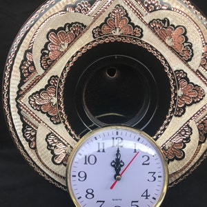 May include: A white clock face with black numbers and hands, set against a black background. The clock is mounted on a decorative copper plate with intricate floral designs.