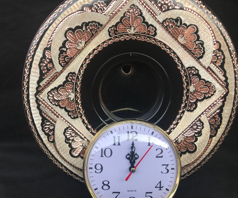 May include: A white clock face with black numbers and hands, set against a black background. The clock is mounted on a decorative copper plate with intricate floral designs.