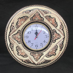 May include: A round, black and white wall clock with copper accents. The clock face has a white background with black numbers and hands. The clock is surrounded by a decorative frame with intricate floral designs.
