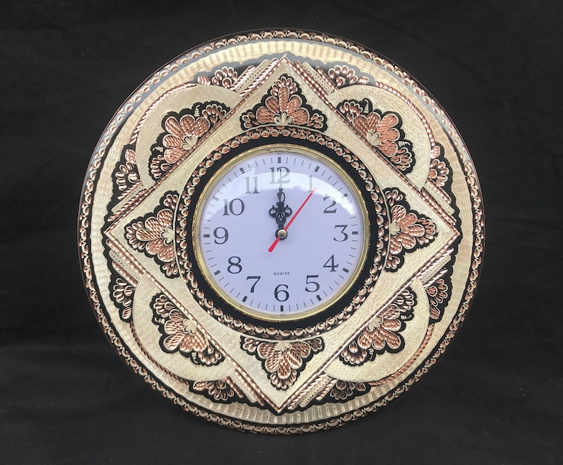 May include: A round, black and white wall clock with copper accents. The clock face has a white background with black numbers and hands. The clock is surrounded by a decorative frame with intricate floral designs.