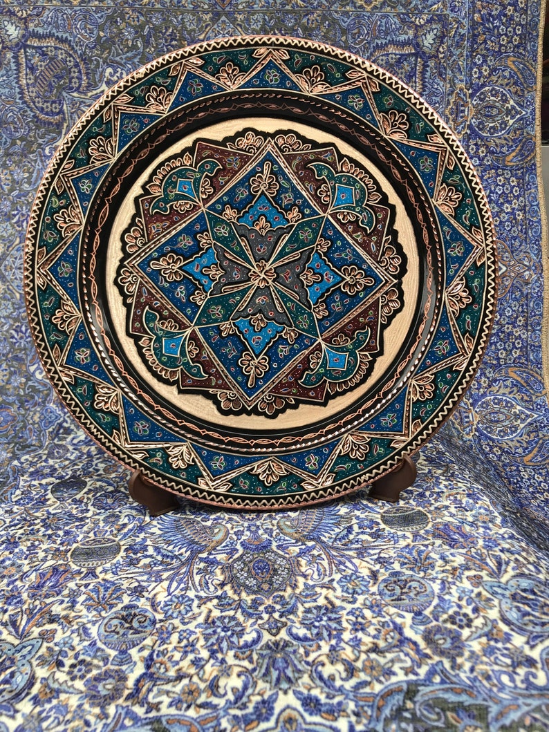 May include: A round copper plate with intricate blue, green, and gold geometric designs. The plate is on a stand and has a black background.