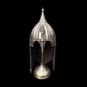 Op de afbeelding: Een antieke messingkleurige helm met ingewikkelde gegraveerde patronen en een maliënkolder nekbescherming. De helm wordt tentoongesteld op een bijpassende standaard tegen een zwarte achtergrond, wat het historische ontwerp en vakmanschap benadrukt.