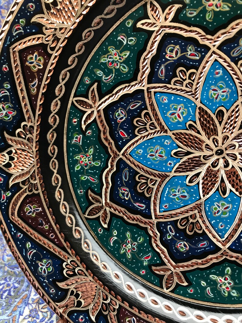 Puede incluir: Un plato decorativo redondo con intrincados dise&ntilde;os florales en tonos azules, verdes y dorados sobre un fondo negro. El plato tiene un borde de cobre.