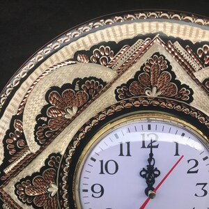 May include: A round, black and white clock with copper accents and intricate floral designs. The clock face is white with black numbers and hands.