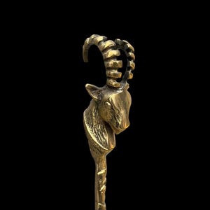 May include: A metallic gold-toned object featuring a sculpted ibex head at the top. The ibex has prominent, curved, ridged horns and detailed facial features. The neck transitions into a slender, textured shaft that extends downward from the head.