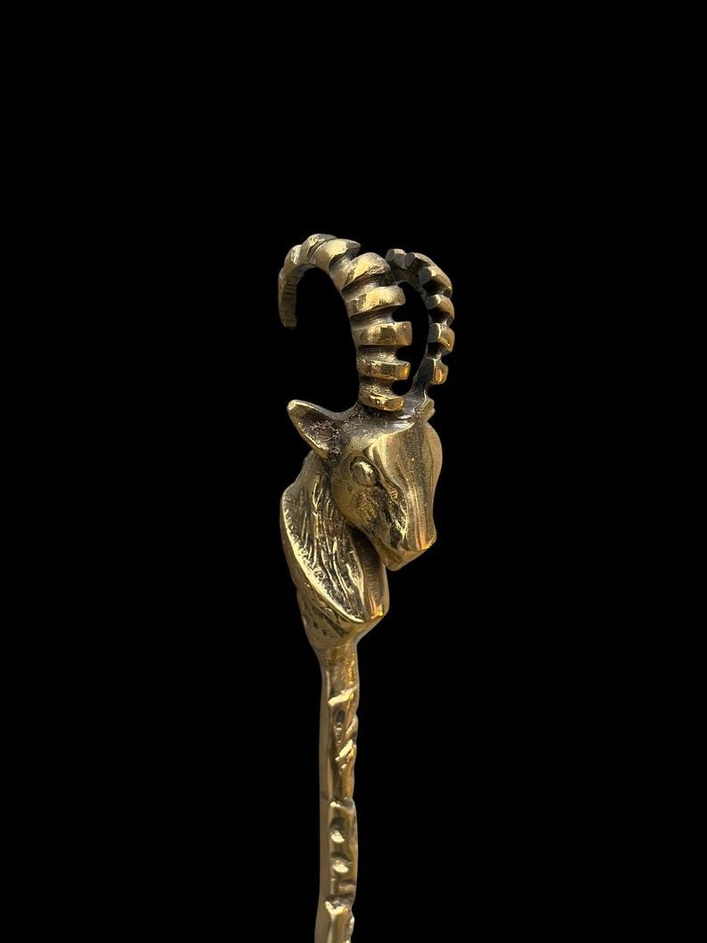 May include: A metallic gold-toned object featuring a sculpted ibex head at the top. The ibex has prominent, curved, ridged horns and detailed facial features. The neck transitions into a slender, textured shaft that extends downward from the head.