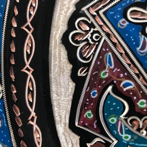 May include: A close-up of a decorative metal plate with intricate designs. The plate features a black background with copper and silver accents. The design includes a floral pattern with a blue, red, and green color scheme.