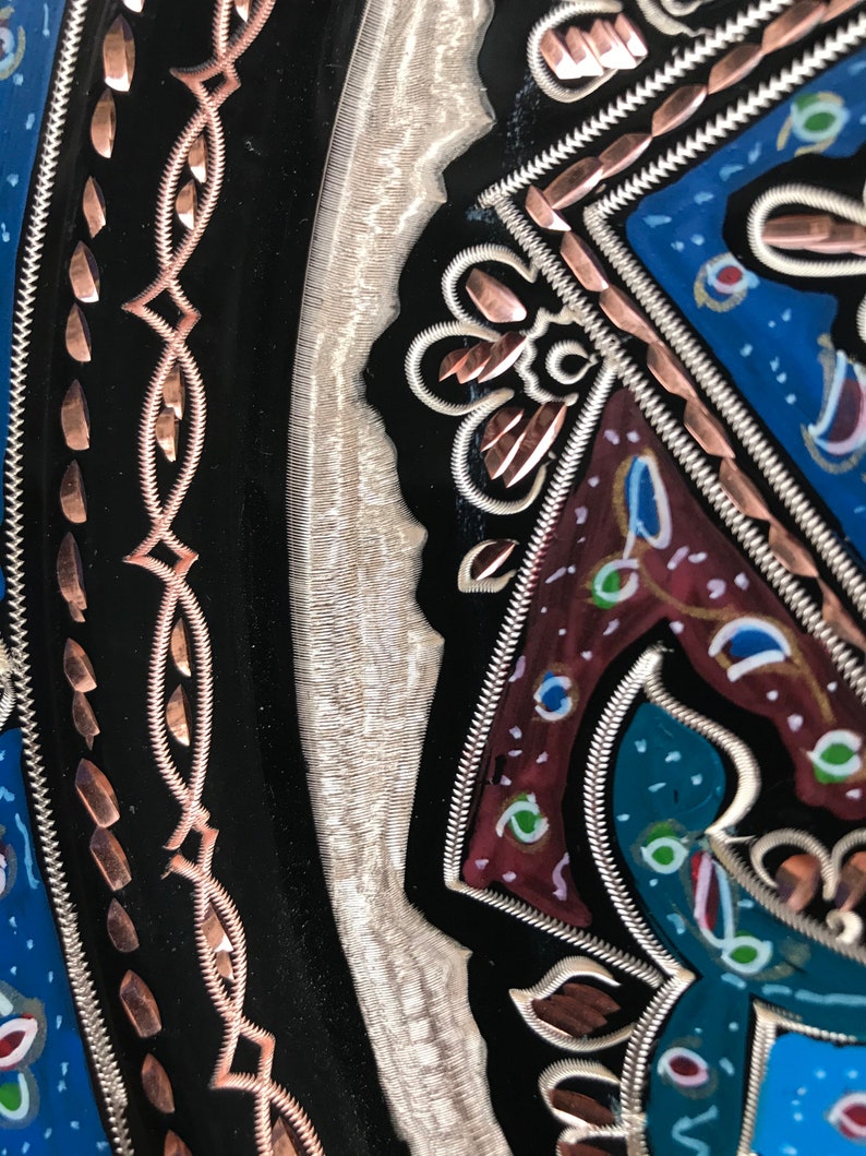 May include: A close-up of a decorative metal plate with intricate designs. The plate features a black background with copper and silver accents. The design includes a floral pattern with a blue, red, and green color scheme.