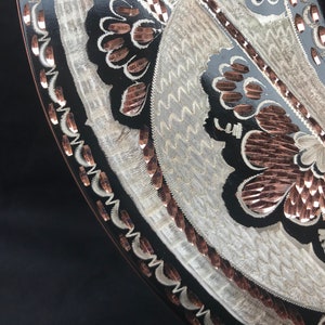 May include: A black circular tray with a silver and copper design. The design features a floral pattern with wavy lines and a border of copper.
