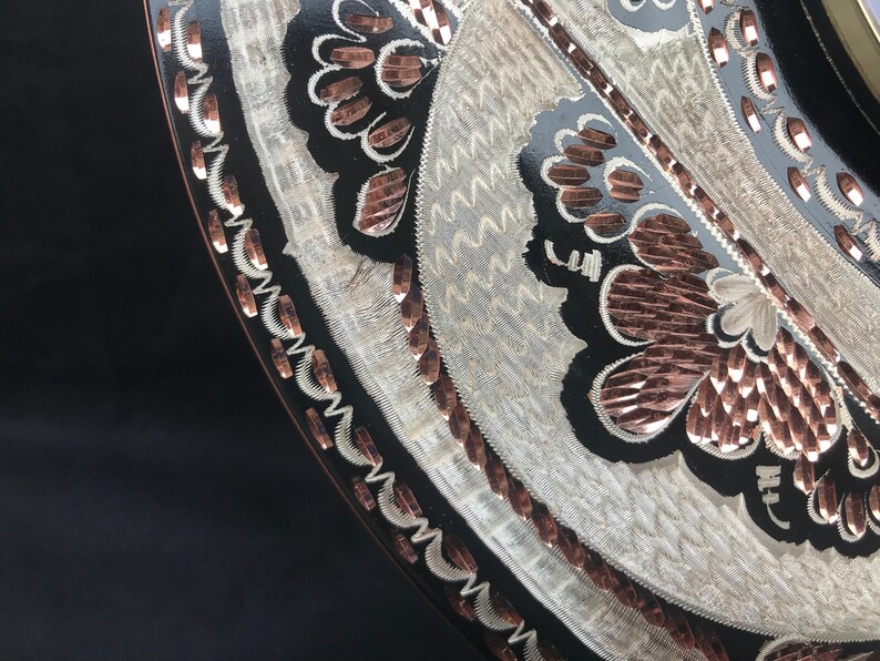 May include: A black circular tray with a silver and copper design. The design features a floral pattern with wavy lines and a border of copper.