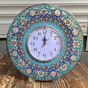 May include: A round wall clock with a decorative face. The clock face is white with black numbers and hands. The outer rim is adorned with a detailed blue, teal, and red floral pattern. The clock is approximately 25 cm in diameter.