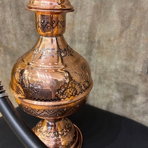 Op de afbeelding: Een koperen waterpijp met ingewikkelde zwart ge&euml;tste ontwerpen. De waterpijp heeft een bolvormig lichaam, een slanke hals en een decoratieve bovenkant. Een zwarte slang met een zilveren spiraal is bevestigd. De waterpijp staat op een zwart oppervlak.