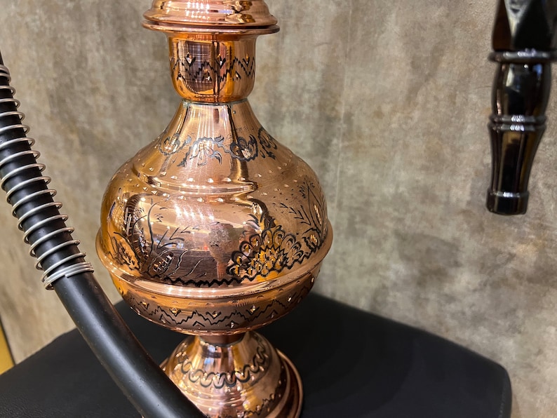 Op de afbeelding: Een koperen waterpijp met ingewikkelde zwart ge&euml;tste ontwerpen. De waterpijp heeft een bolvormig lichaam, een slanke hals en een decoratieve bovenkant. Een zwarte slang met een zilveren spiraal is bevestigd. De waterpijp staat op een zwart oppervlak.