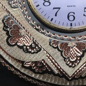 May include: A black clock with a white face and gold trim. The clock has a decorative design with copper and silver accents. The clock face reads "quartz" and has numbers 4, 5, 6, 7, and 8.