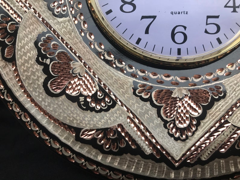 May include: A black clock with a white face and gold trim. The clock has a decorative design with copper and silver accents. The clock face reads "quartz" and has numbers 4, 5, 6, 7, and 8.