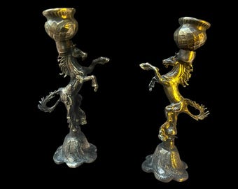 May include: Two decorative candle holders with a horse design. One is silver-toned, the other gold-toned. Each features a rearing horse supporting a candle cup. The bases have a textured, organic shape. The background is black.