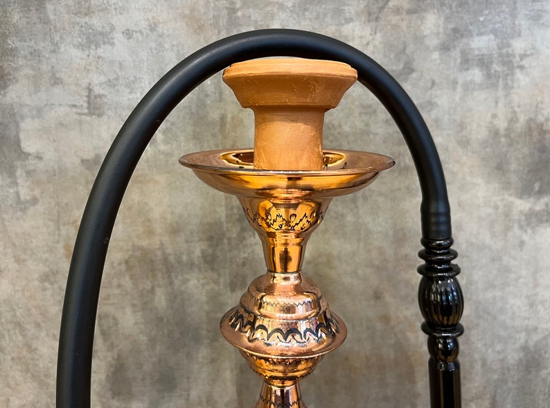Op de afbeelding: Een koperen waterpijp met een zwarte slang en een kleikom. De waterpijp heeft een sierlijk ontwerp op de basis en een decoratieve steel. De slang buigt over de bovenkant van de waterpijp. De achtergrond is een getextureerde grijze muur.