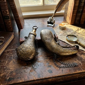 Op de afbeelding: Twee antieke messing poederflessen met ingewikkelde gegraveerde ontwerpen. Eén is een ronde fles met een ketting, de andere is hoornvormig. Ze liggen op een houten oppervlak met oude boeken en een kaart.