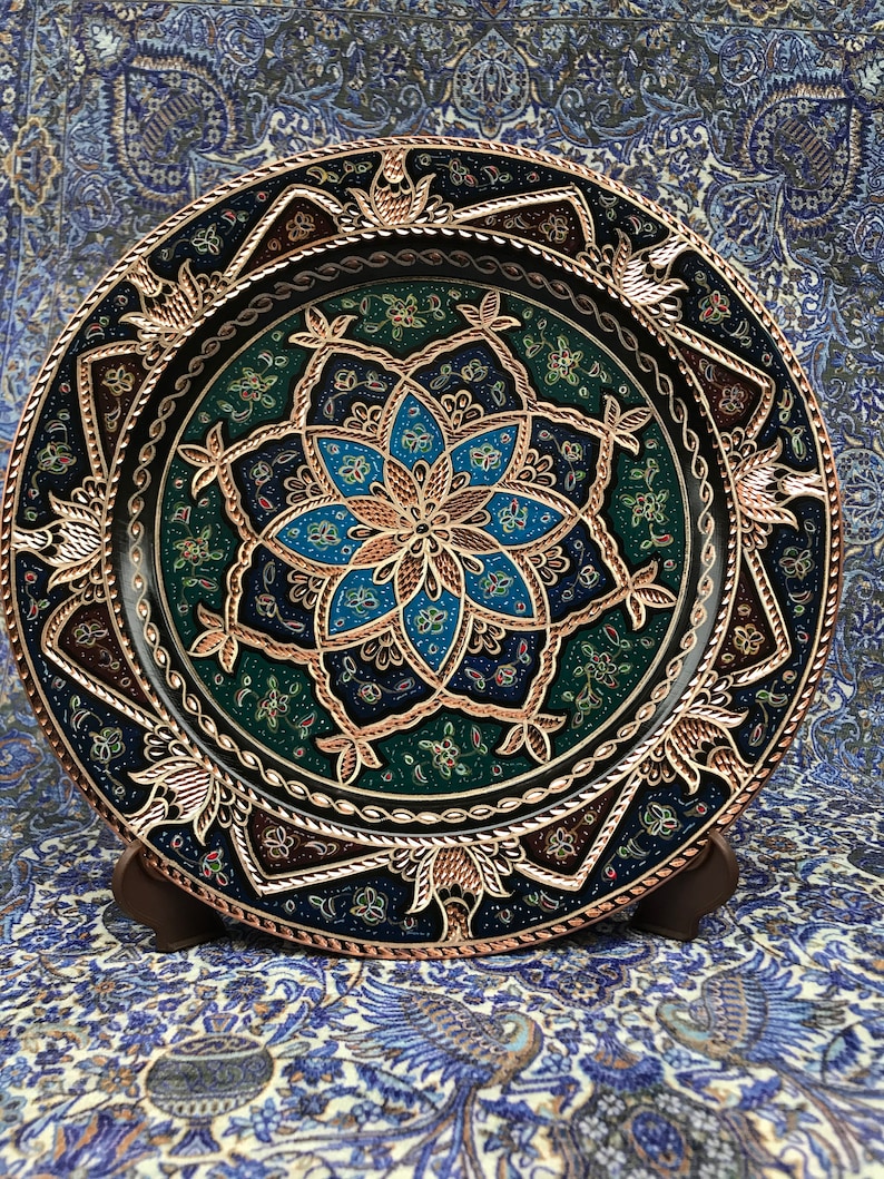 Puede incluir: Un plato redondo decorativo con un dise&ntilde;o geom&eacute;trico azul, verde y dorado. El plato est&aacute; hecho de metal y tiene un borde elevado.